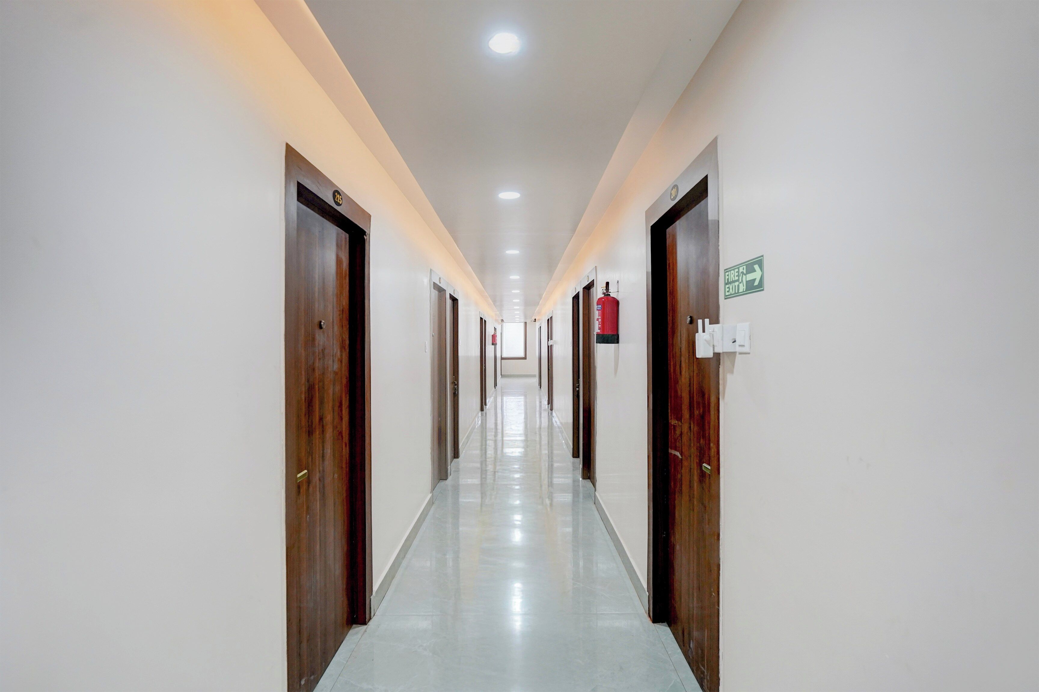 Un largo y estrecho pasillo de hotel con puertas a ambos lados, un letrero de salida de emergencia y un extintor colgado en la pared.