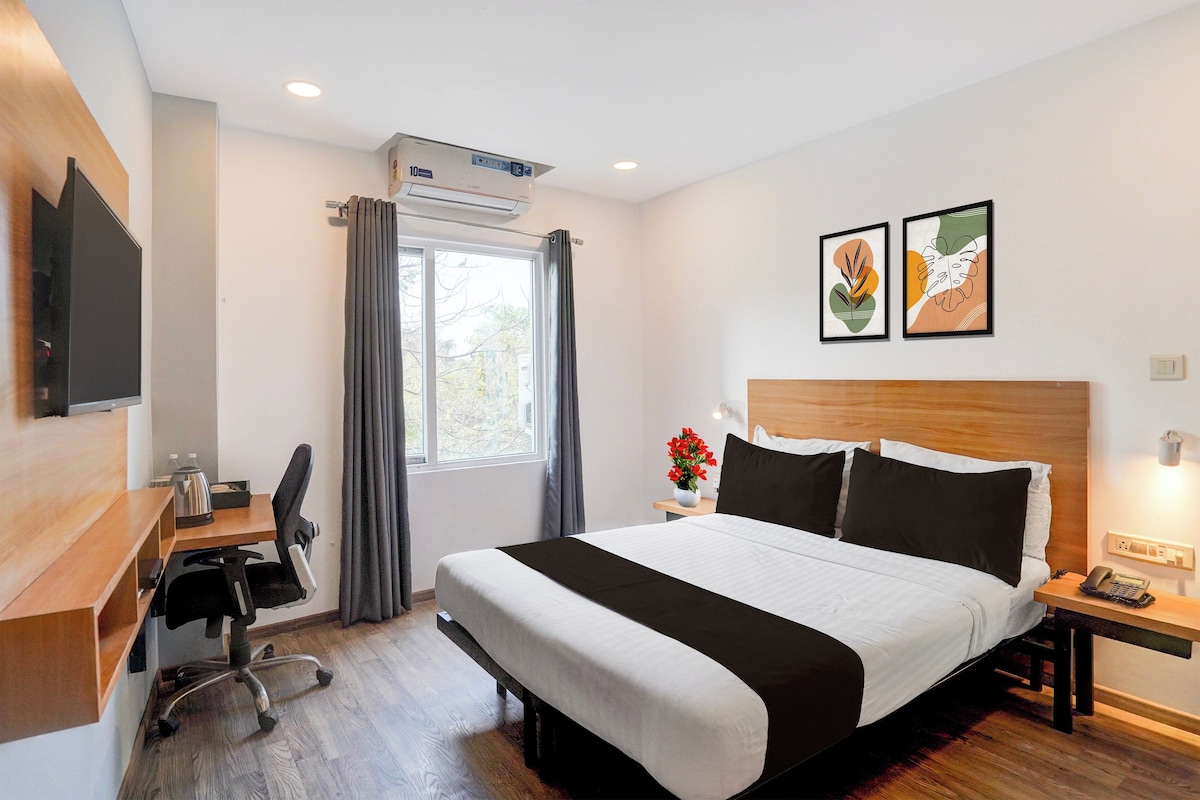 A hotel room with a bed, a desk, a chair, a TV, a window with curtains, and two framed pictures on the wall.