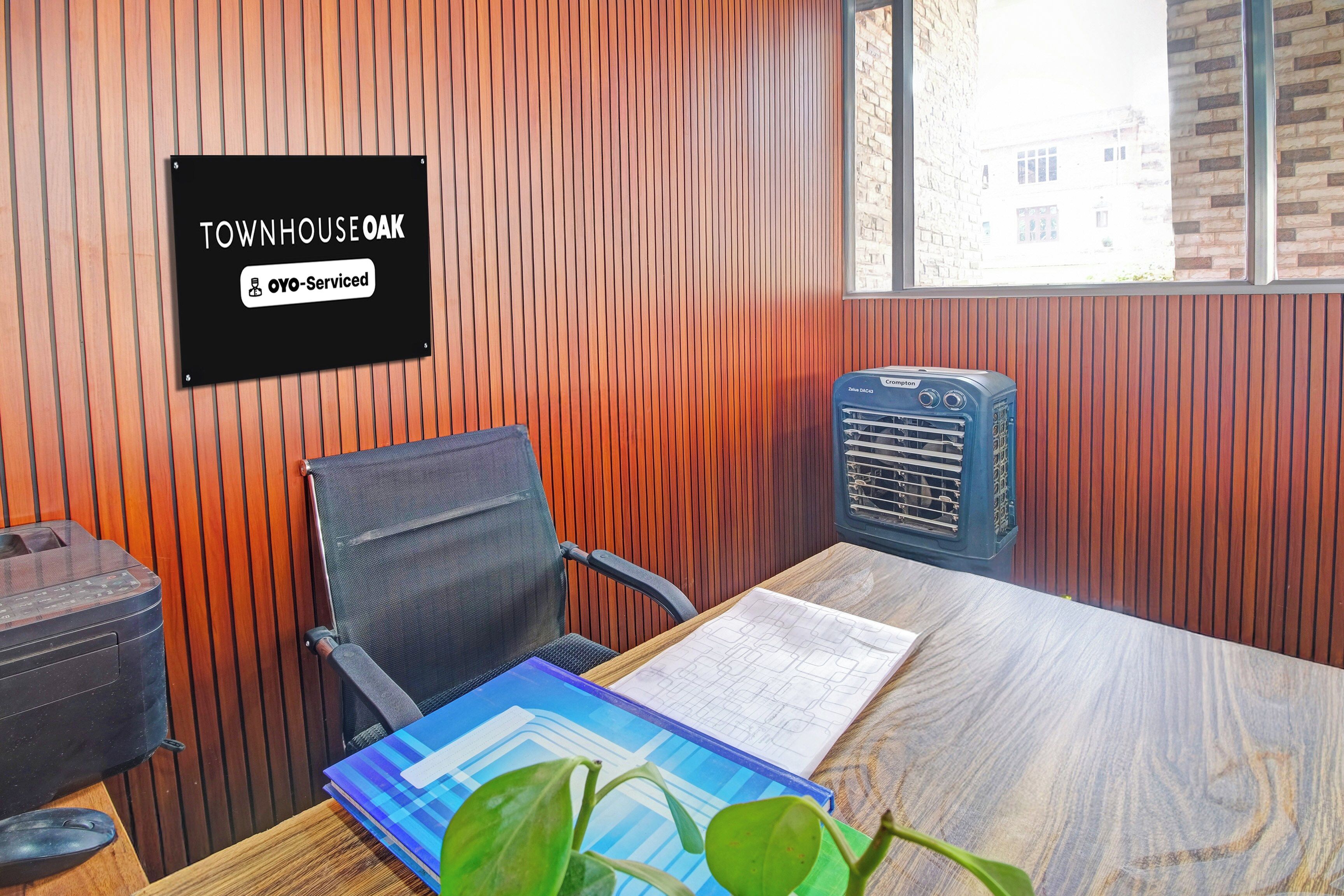 Een kamer met een houten wandpaneel, een zwart bord met de tekst 'TOWNHOUSE OAK OYO-Serviced', een blauwe airconditioner, een zwarte bureaustoel, een houten tafel met een plant en een notitieboek.