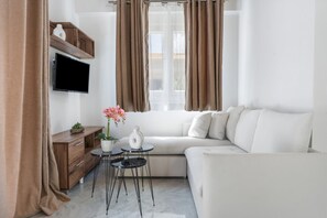 Ein modernes Wohnzimmer mit einer Couch, einem kleinen Tisch, einem an der Wand montierten Fernseher und einem Fenster mit Vorhängen.