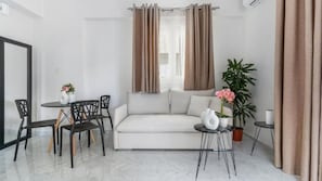A modern living room with a white sofa, a dining table with chairs, a potted plant, and a small side table. - Maria Ground Floor - Miralis Apartments (Episkopos)