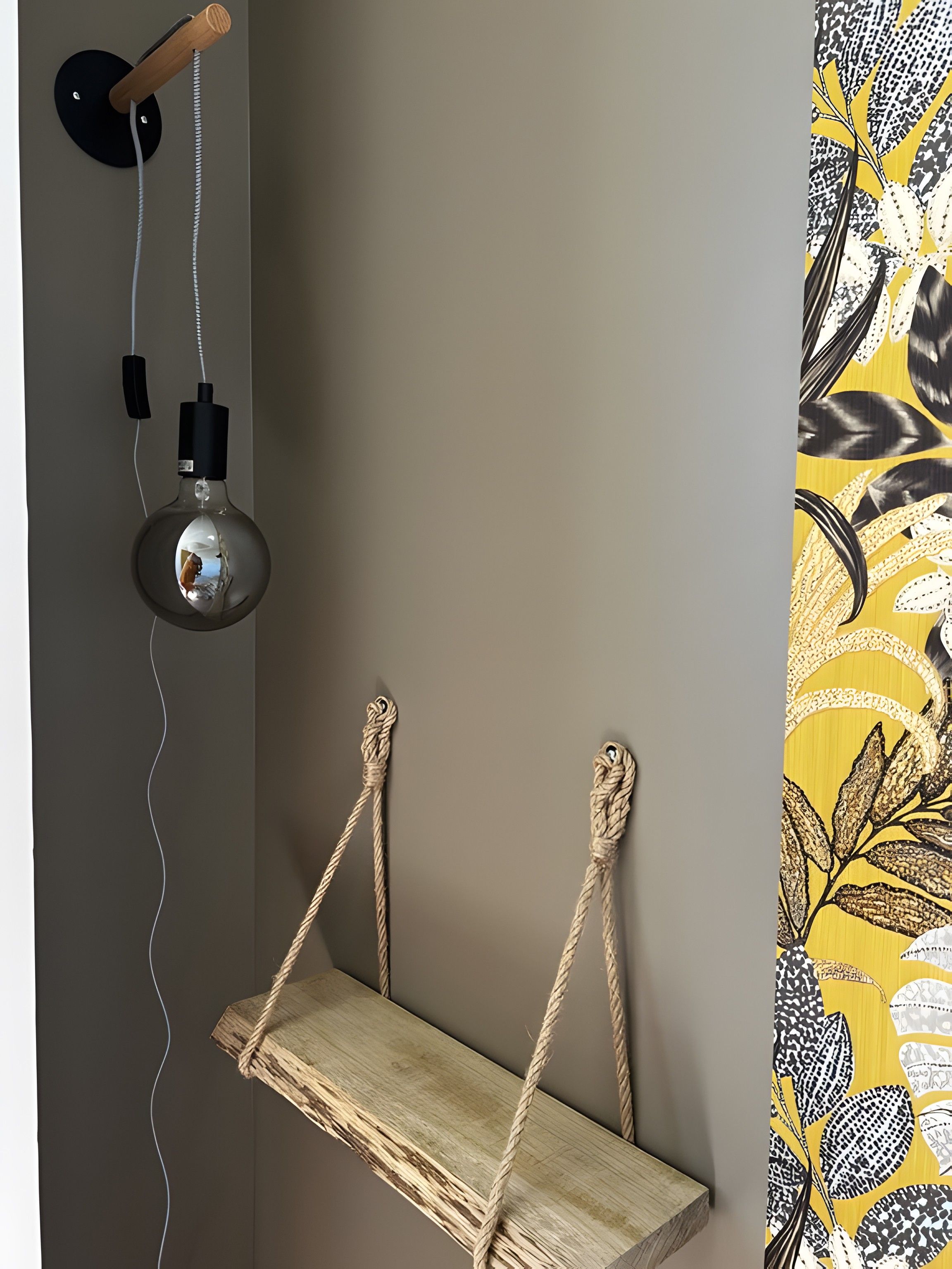 A room with a wooden shelf suspended by ropes, a wall-mounted lamp, and a patterned curtain.