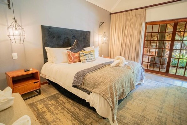 A bedroom with a large bed, a nightstand, a wooden side table, and a view of greenery through a sliding glass door.