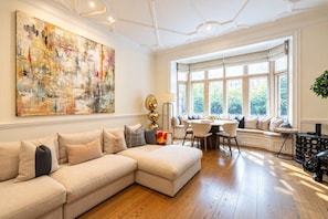 A spacious living room with a large abstract painting, a beige sectional sofa, a dining area with a round table, and a view through large windows. - Grand Knightsbridge Home High Ceilings Easy Access (London)