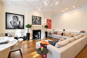 A modern living room with a fireplace, a sofa, a dining table with chairs, and artwork on the walls. - Grand Knightsbridge Home High Ceilings Easy Access (London)
