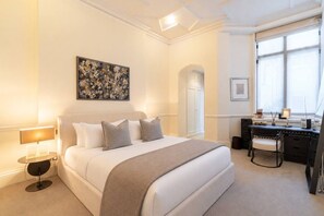 A hotel room with a large bed, a desk, a chair, a lamp, and a window. - Grand Knightsbridge Home High Ceilings Easy Access (London)