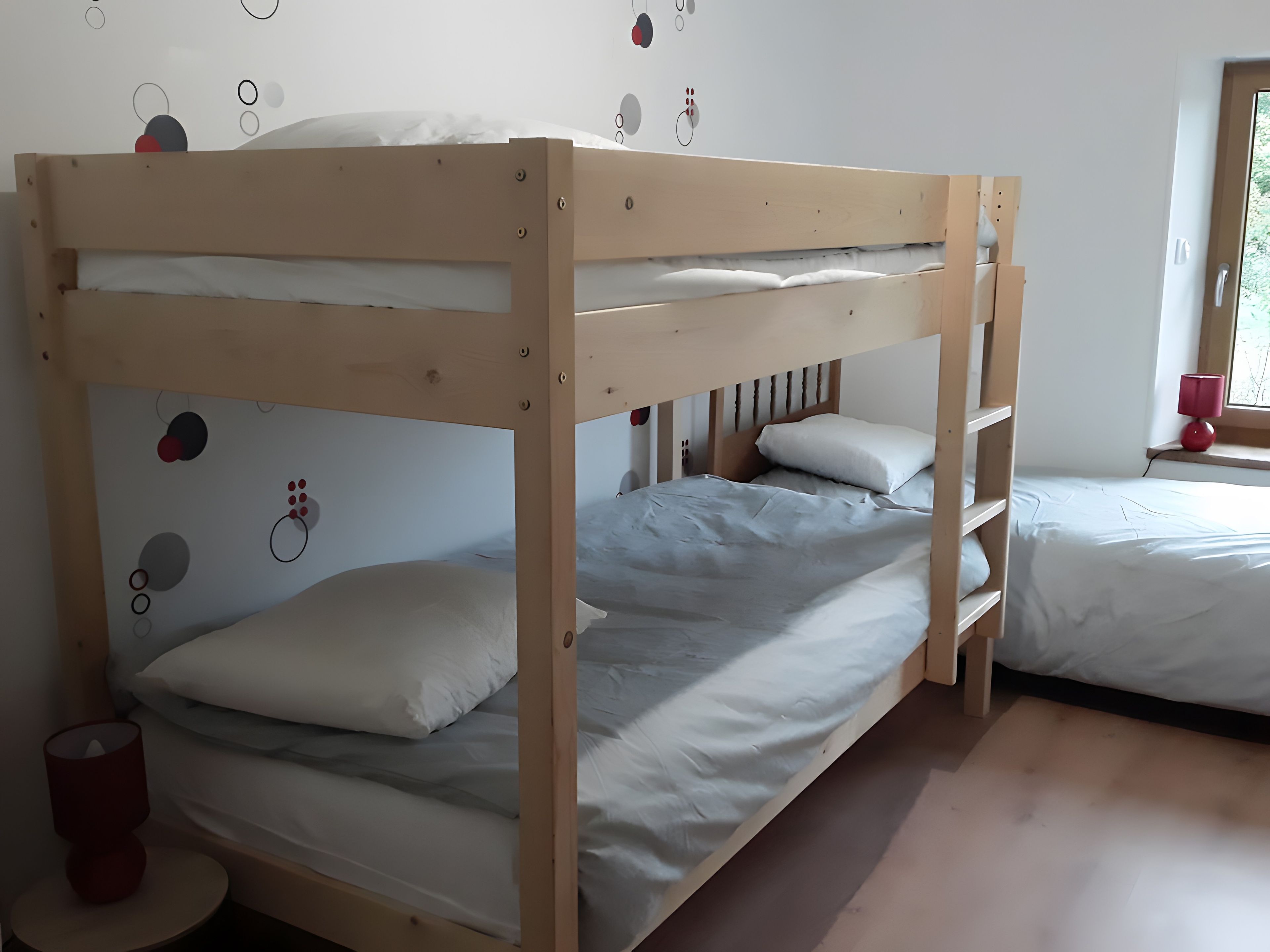 A bunk bed room with a wooden frame, white bedding, and a red lamp on a bedside table.