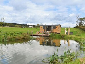 A wooden cabin with a dock extending over a calm pond, surrounded by lush greenery and a clear sky. - Shepherd Hut at Garden Cottage (Chepstow)