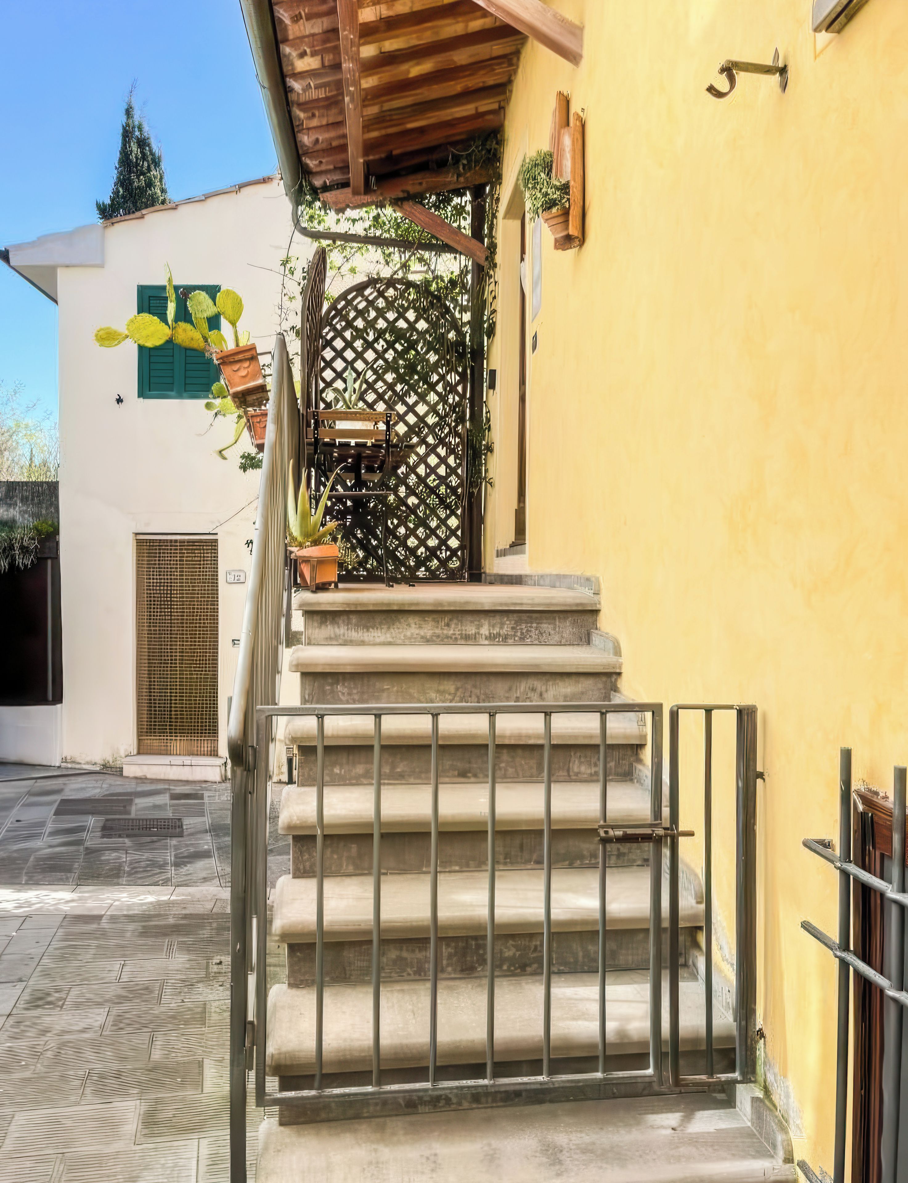Un escalier menant à un bâtiment jaune, doté d’une grille en métal, de plantes en pot et d’un plafond en bois.