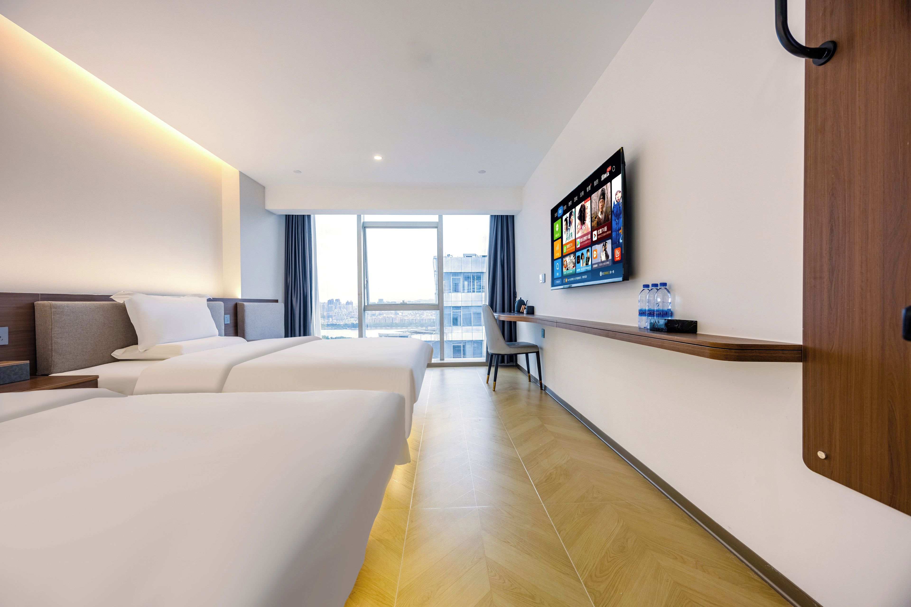 Een hotelkamer met twee bedden, een aan de muur bevestigde televisie, een bureau met stoel en uitzicht op de stad door grote ramen.