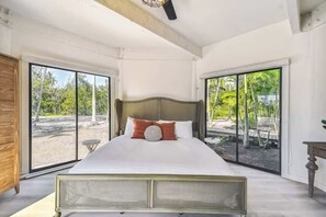 Ein großes Bett mit Kopfstütze, ein Nachttisch aus Holz und Blick durch eine Glastür auf einen Außenbereich.