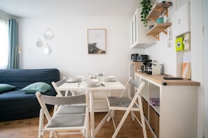 A modern living room with a sofa, dining table, and chairs. There is a kitchen area with appliances and a plant. - L’Écrin d’Ax - 500 mètres des télécabines (Ax-les-Thermes)