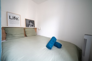 A neatly made bed with green bedding and two rolled blue towels, a framed picture of a trumpet player, and a white air conditioning unit. - L’Écrin d’Ax - 500 mètres des télécabines (Ax-les-Thermes)