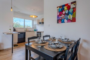 A modern kitchen with a dining area, featuring a large window, a dishwasher, and a dining table set with plates, glasses, and a candle. - Les Caraïbes Bretons - 500m de la plage (Saint-Pabu)