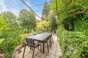 Une terrasse extérieure avec une table et des chaises, entourée d’une végétation luxuriante et d’arbres.