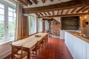A dining area with a wooden table and chairs, a brick wall, and a kitchen with a sink. - Le Charme et le Chevreuil - Maison Normande (Rosay)