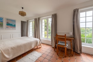 A bedroom with a bed, a desk, and a chair. There are two windows with curtains, a painting on the wall, and a hanging light fixture. - Le Charme et le Chevreuil - Maison Normande (Rosay)