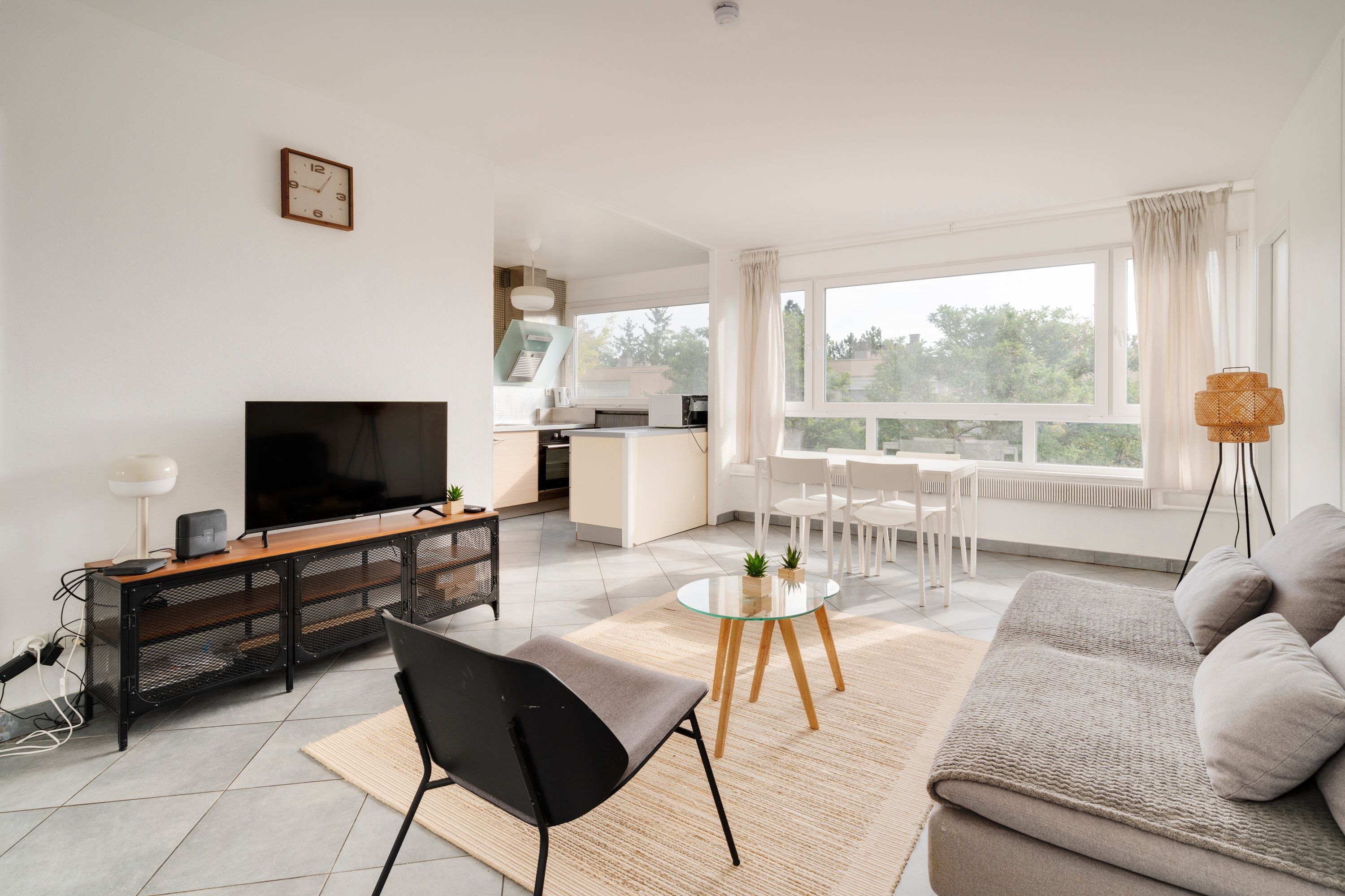 A modern living room with a flat-screen TV, a sofa, a dining table with chairs, and a kitchen in the background.
