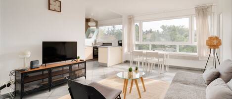 A modern living room with a flat-screen TV, a sofa, a dining table with chairs, and a kitchen in the background.