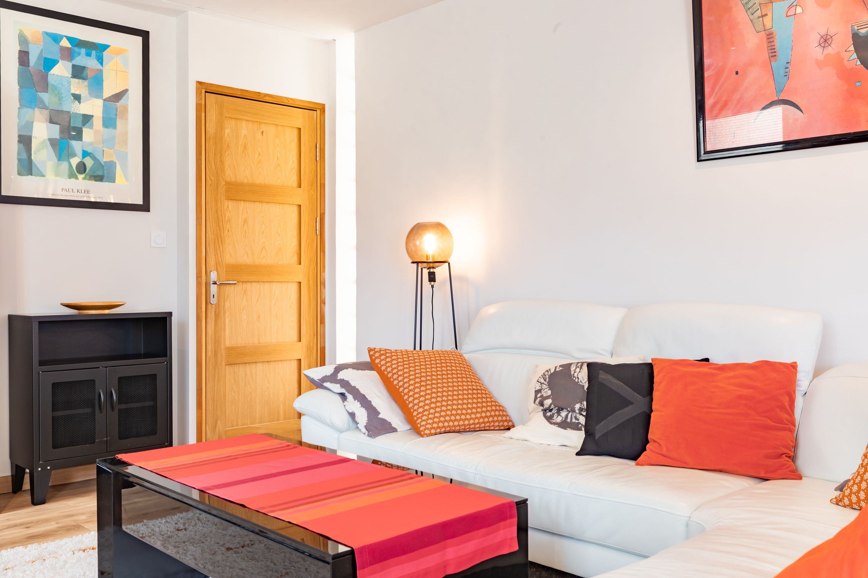A modern living room with a white sofa, colorful cushions, a coffee table, a lamp, and artwork on the walls.