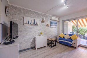 A living room with a white brick wall, a flat-screen TV, a white mini-fridge, a blue sofa with yellow cushions, and a table with a vase of flowers. - Launa - 2 min à pied de la mer (Roquebrune-sur-Argens)