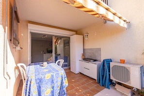 A patio area with a table covered in a floral tablecloth, a white chair, a sliding glass door leading to an indoor space, and a white cabinet with a television. - Launa - 2 min à pied de la mer (Roquebrune-sur-Argens)