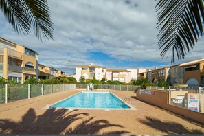 A swimming pool surrounded by a fenced area with palm trees, set against a backdrop of multi-story buildings. - Le Cocon du Cap d'Agde - 5 min du port (Agde)