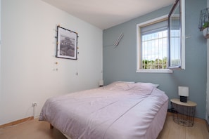 A bedroom with a bed, a window, a bedside table, and a wall-mounted lamp. - Le Cocon du Cap d'Agde - 5 min du port (Agde)