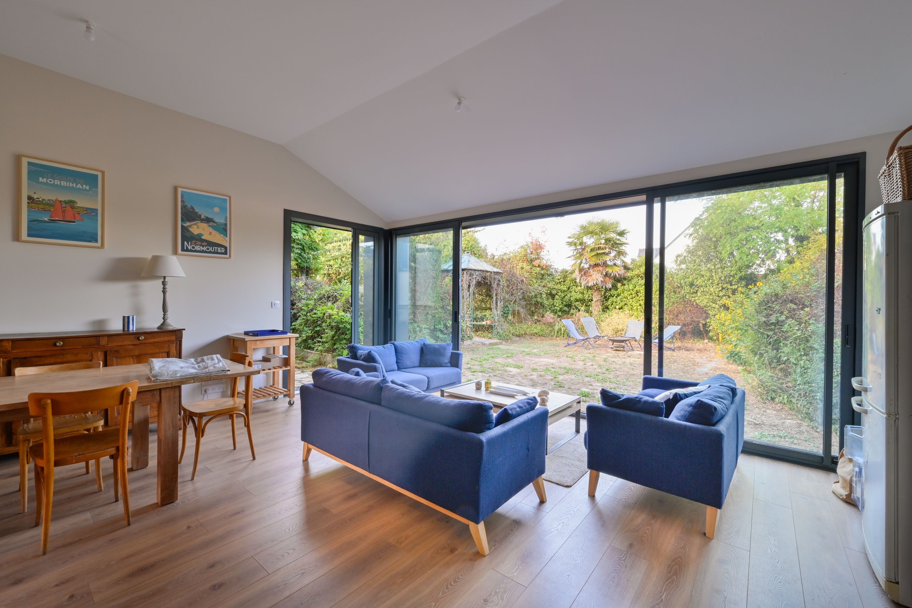 Un salon moderne avec une grande fenêtre, un coin repas avec des meubles en bois et deux canapés bleus.