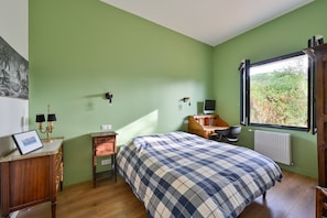 A bedroom with a bed, bedside tables, a desk, a chair, a radiator, and a window with a view of greenery. - La Maison du Logeo - A 300 m du Golfe (Sarzeau)