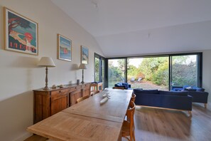 Un coin repas avec une table en bois, des chaises et un canapé. On y trouve deux lampes fixées au mur et des tableaux encadrés. La pièce est dotée de grandes fenêtres qui offrent une vue sur la verdure environnante.