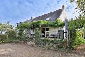 A house with a sloped roof, a visible chimney, and a front porch with outdoor seating. - La Maison du Logeo - A 300 m du Golfe (Sarzeau)