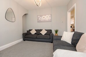 A living room with a grey sofa, a smaller grey sofa, a mirror, a wall-mounted picture, and a doorway leading to another room. - Toast Stays - Teal View House (Darlington)