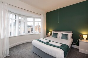 Et hotelværelse med en seng, et natbord, en lampe og udsigt ud af vinduet med persienner.