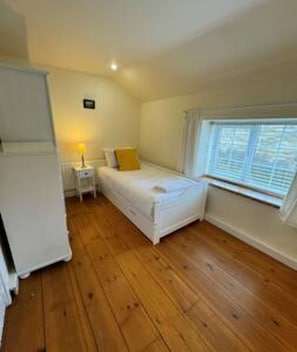 A small, single-bed room with a wooden floor, a bedside table with a lamp, and a window with blinds. - Toast Stays - Woodbine Cottage (Alston Moor)