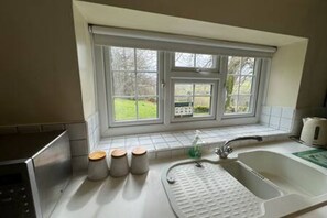 A kitchen with a window, a sink, a kettle, and a microwave. - Toast Stays - Woodbine Cottage (Alston Moor)