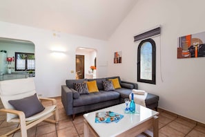 A living room with a grey sofa, a wooden chair, a coffee table, and a window with a view. - Blue Beach - Three Bedroom Villa, Sleeps 6 (Tías)