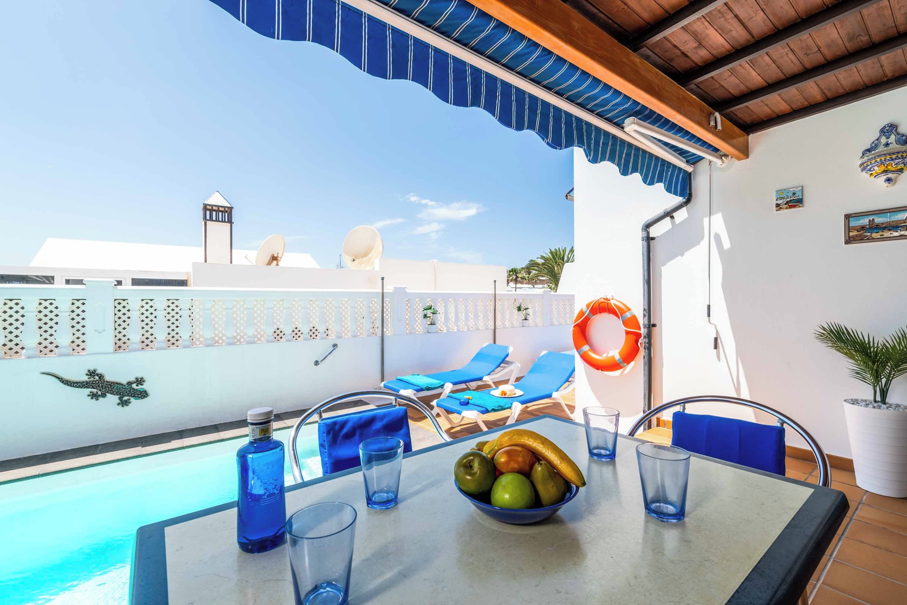 Un espace aménagé au bord de la piscine, avec des chaises longues, une table garnie de fruits et de boissons, et un anneau de sauvetage accroché au mur.