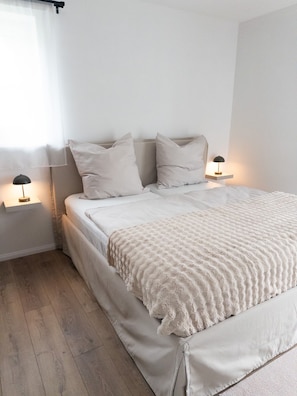 A bedroom with a bed, two bedside tables, a window with curtains, and a wooden floor.