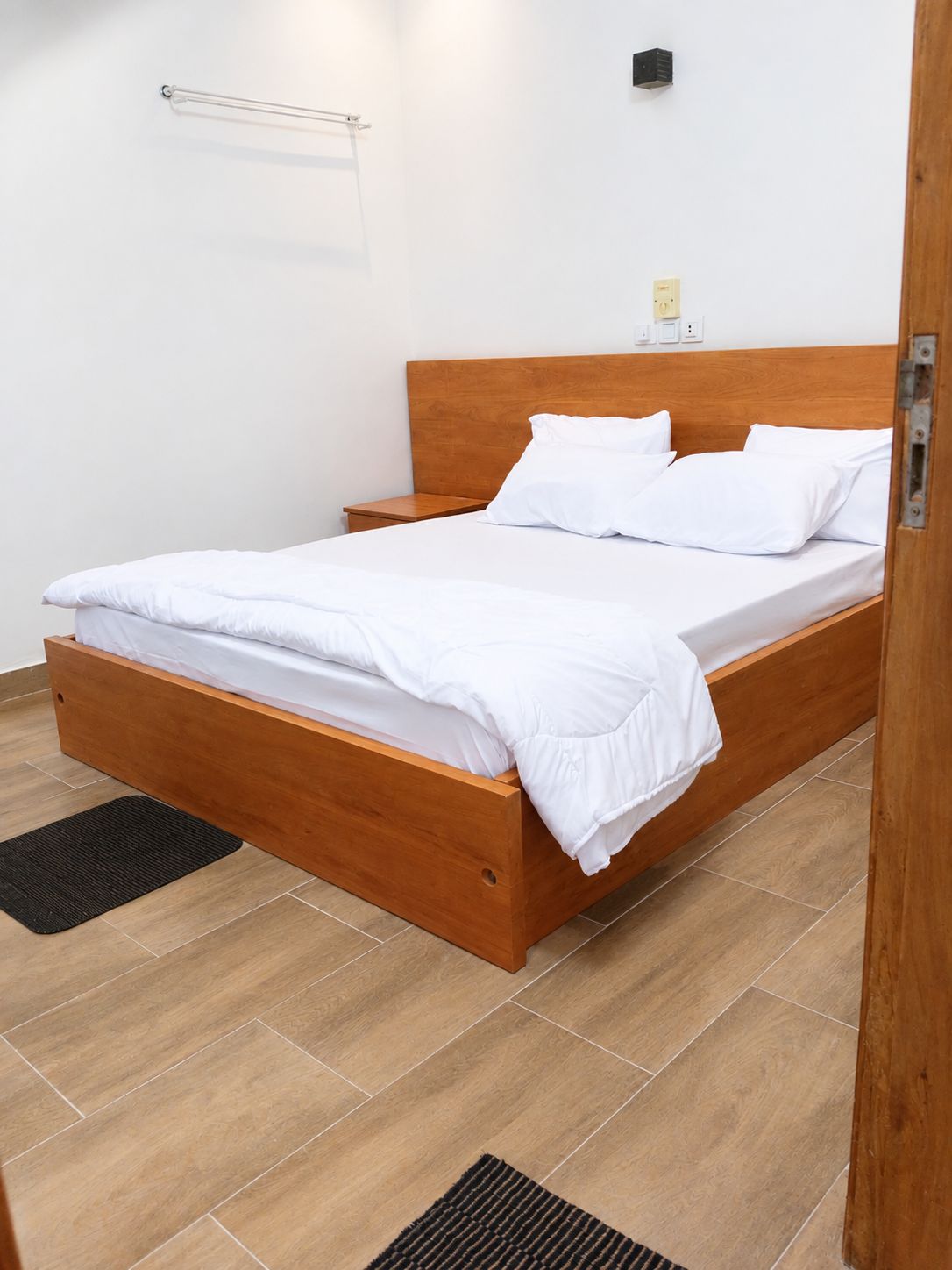 Una cama individual con estructura de madera y ropa blanca en una habitación con suelo de baldosas.