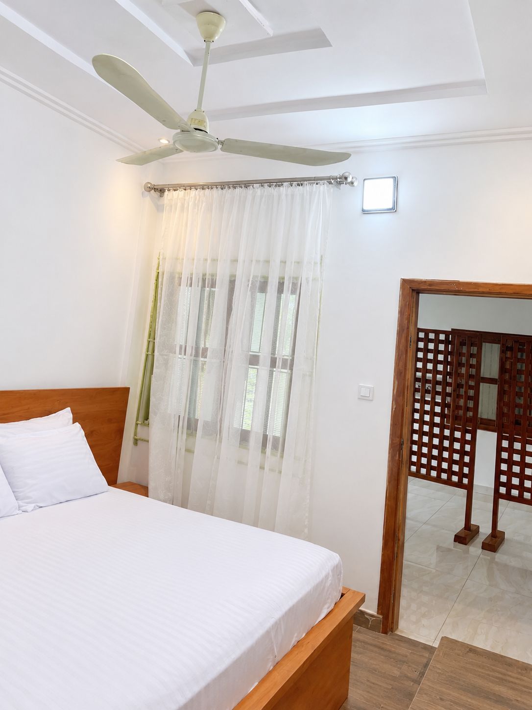 Une chambre à coucher comprenant un lit, un ventilateur de plafond, une fenêtre avec des rideaux légers et une cloison en bois.