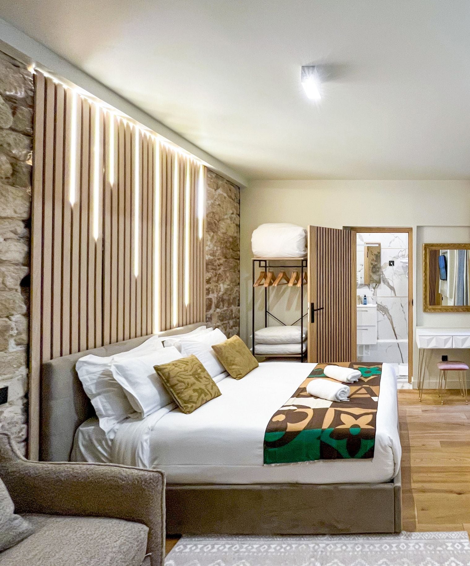 A modern hotel room with a large bed, a seating area, a bathroom with a mirror, and a stone wall.