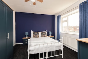 A bedroom with a white metal bed frame, a dark blue wall, a window with curtains, and a ceiling fan. - Toast Stays - The Pittington Loft (High Pittington)
