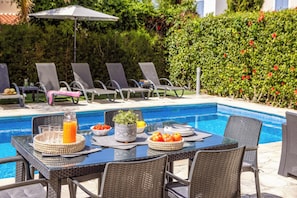 Un espace repas aménagé au bord de la piscine, avec une table dressée pour deux personnes, entourée de chaises longues, d'une petite table sur laquelle se trouve une carafe de jus d'orange, un saladier de tomates, une plante en pot et un panier de pommes.