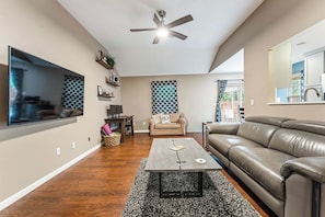 A living room with a flat-screen TV, a ceiling fan, a sofa, a coffee table, and a patterned rug. - Riders Ready! Pet Friendly Downtown Bentonville/Momentary Location (Bentonville)