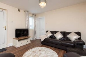 A living room with a dark leather sofa, a flat-screen TV on a wooden stand, a round white rug, and a chandelier. - Toast Stays - The Langley Nook (Hill Top)