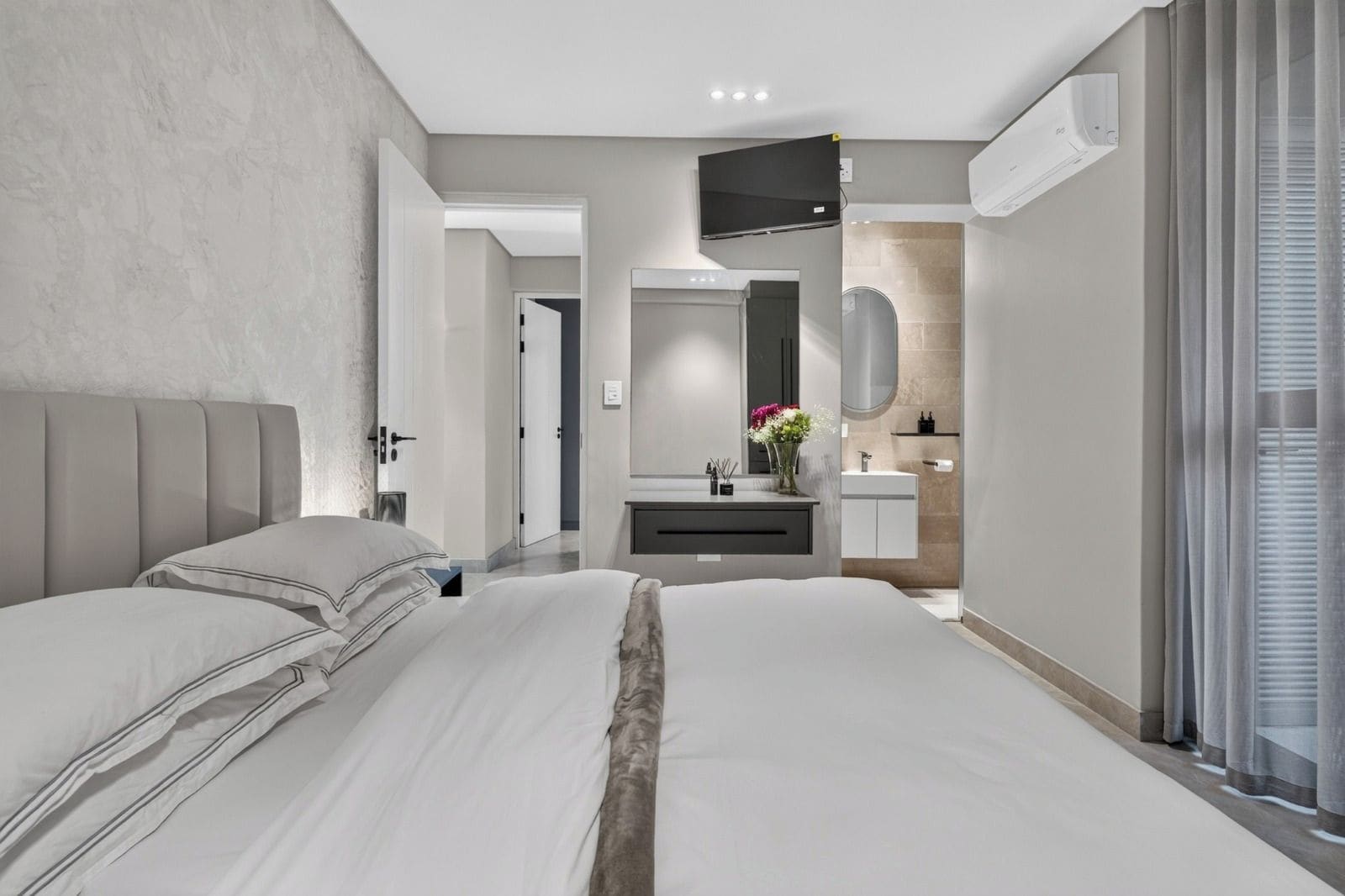 Un dormitorio moderno con una cama grande, una mesita de noche, televisión integrada y un baño con espejo y lavamanos.