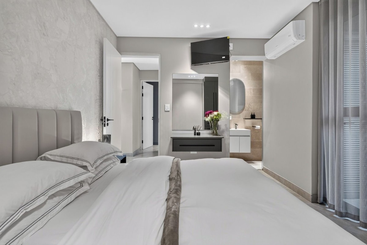 A modern bedroom with a large bed, a bedside table, a built-in TV, and a bathroom with a mirror and sink.