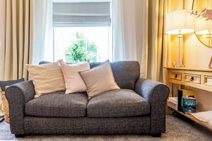 A grey sofa with multiple cushions, a window with curtains, a lamp, and a bookshelf with books. - Toast Stays - Pleasant View House (Crook)