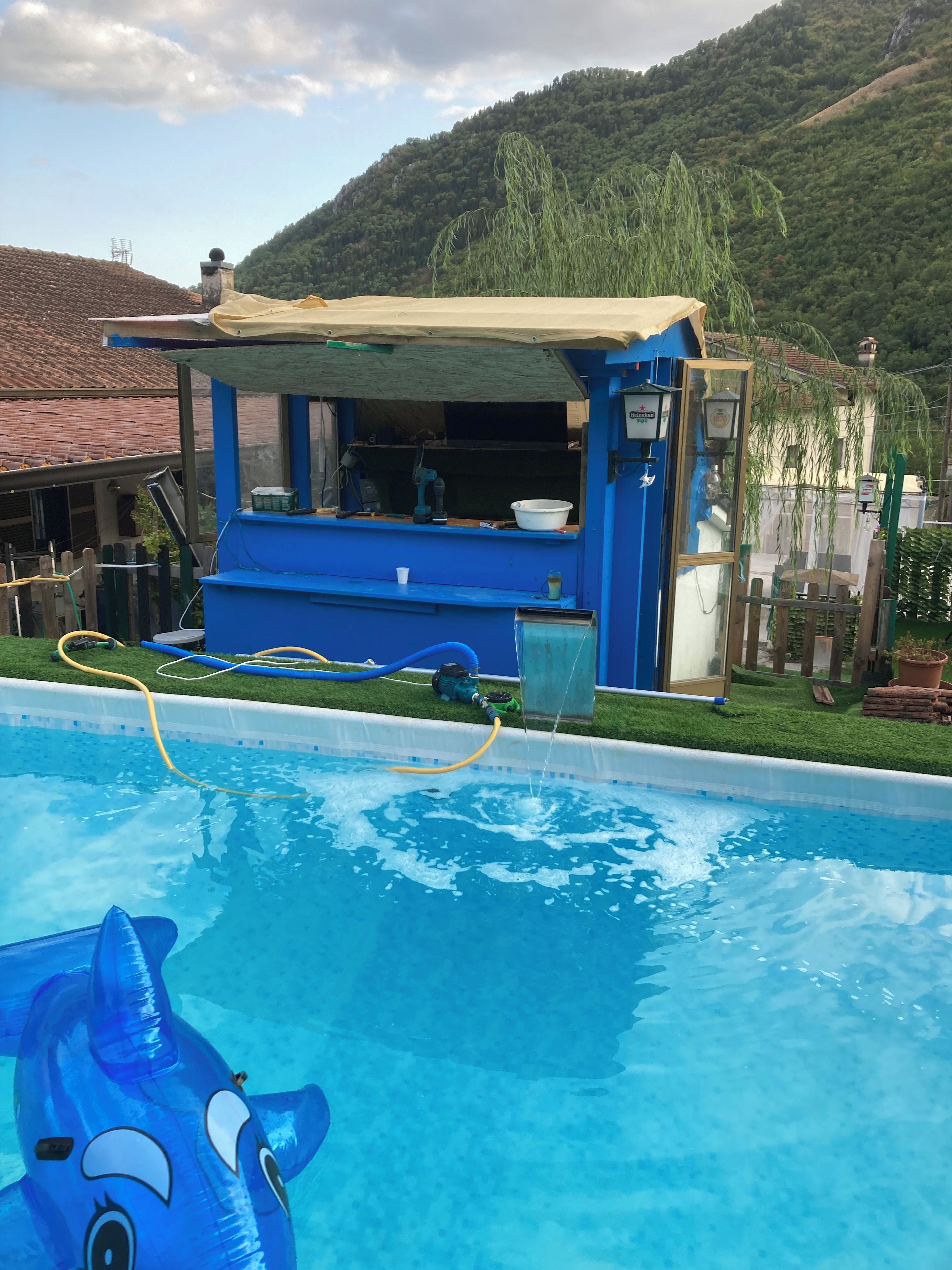 Une piscine avec un jouet en forme de requin gonflable bleu, une structure bleue et beige dotée d’une fenêtre, et une colline verdoyante en arrière-plan.